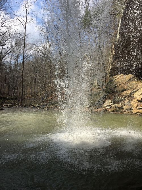 A water fountain erupts from a rocky area, surrounded by trees and a calm river under a partly cloudy sky.