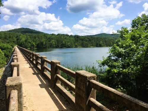 A scenic view of a bridge over a calm lake, surrounded by lush greenery and mountains under a partly cloudy sky.