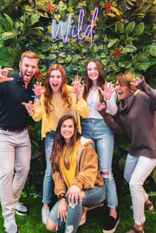 A group of five people poses playfully in front of a green wall with the word "Wild" in neon lights.