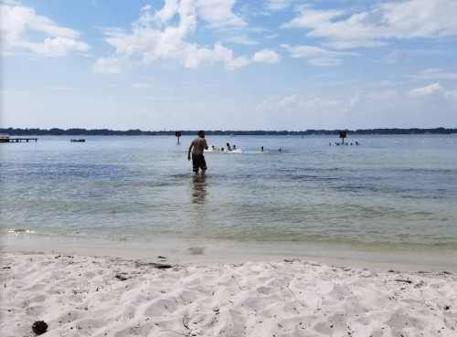 A person wades into a calm lake, with others swimming nearby and a sandy beach in the foreground under a blue sky.