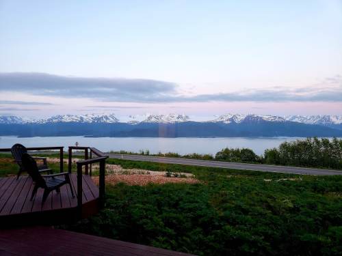 A scenic view of snow-capped mountains and a calm lake, with a wooden deck and chairs in the foreground.