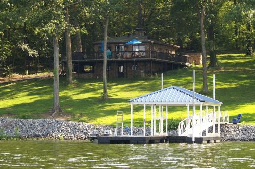 A lakeside view featuring a wooden house on a hill and a small dock with a boat lift in the foreground.