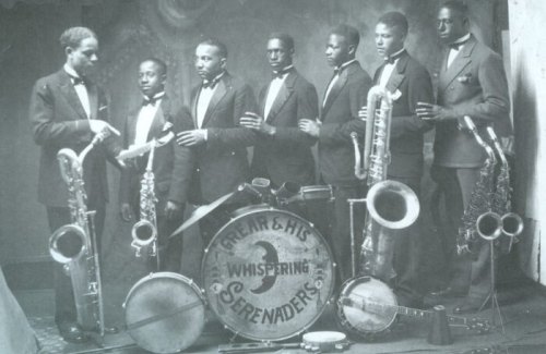 A vintage black-and-white photo of seven musicians in suits, posing with various instruments, including saxophones and a drum.
