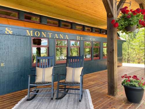 Two blue rocking chairs with cushions sit on a wooden porch, surrounded by potted flowers and a vintage train car backdrop.