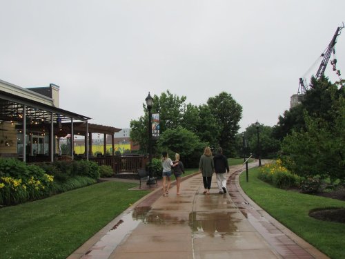 Four people walk along a wet pathway beside a restaurant and greenery on a cloudy day.