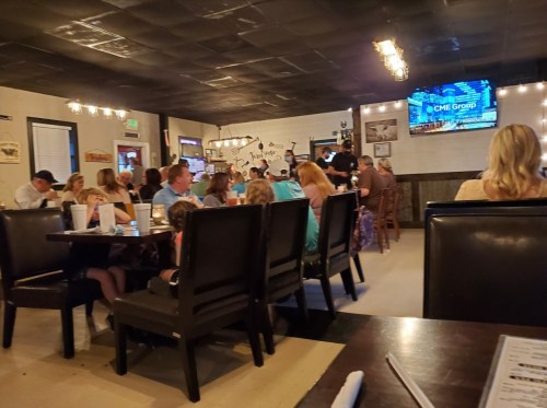 A busy restaurant interior with diners at tables and a TV displaying a business logo on the wall.