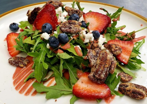 A vibrant salad with arugula, strawberries, blueberries, goat cheese, and candied pecans, drizzled with dressing.