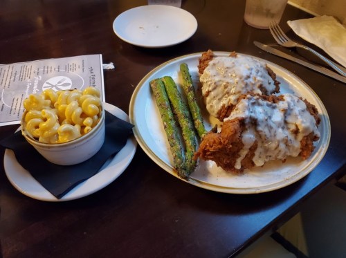 A plate with fried chicken smothered in gravy, asparagus, and a side of macaroni and cheese on a dark wooden table.
