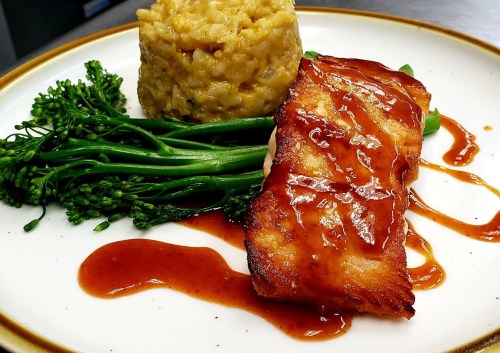 Grilled salmon fillet with a glaze, served with broccolini and a mound of creamy risotto on a white plate.