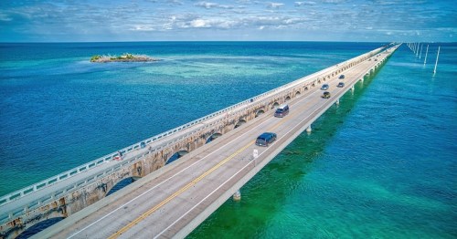 Aerial view of a long bridge stretching over turquoise waters, with cars driving and a small island in the distance.