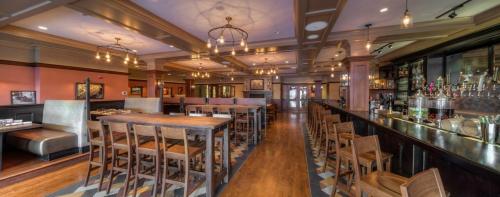 Interior of a restaurant featuring wooden tables, chairs, a bar, and warm lighting, creating a cozy atmosphere.