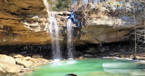 A serene waterfall cascades over rocks into a tranquil green pool, surrounded by winter trees and ice formations.