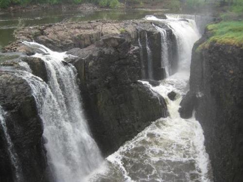 A scenic view of a waterfall cascading over rocky cliffs into a river below, surrounded by lush greenery.