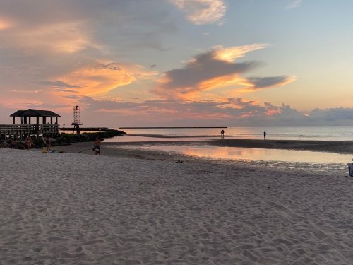 A serene beach at sunset, with colorful clouds, a gazebo, and people walking along the shore.