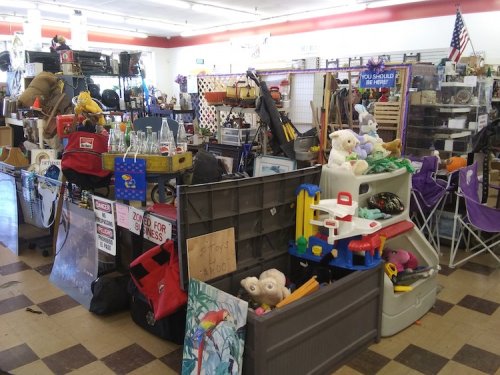 A cluttered thrift store aisle filled with various items, including toys, furniture, and household goods.
