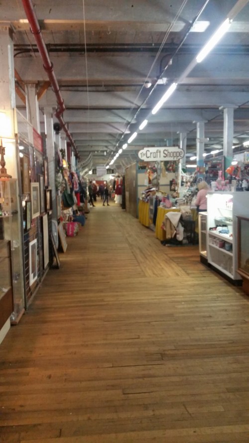 A spacious indoor market with wooden floors, lined with vendor booths and shoppers browsing various items.