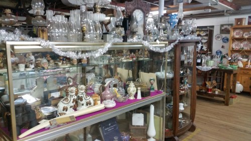 A vintage shop display featuring various antiques, glassware, and decorative items, adorned with tinsel.