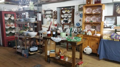 A vintage shop display featuring various antiques, kitchenware, and decorative items on shelves and a wooden cart.