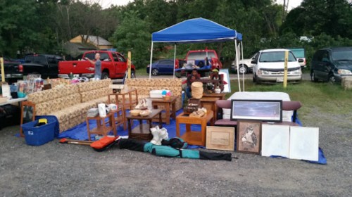 A yard sale setup with a blue tent, furniture, artwork, and various items displayed on a gravel surface.