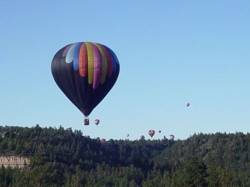 A colorful hot air balloon floats above a forested landscape, with more balloons visible in the distance against a clear sky.
