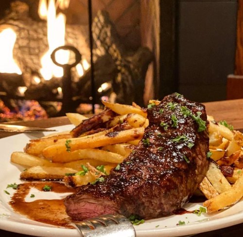A juicy steak with crispy fries, garnished with herbs, served in front of a cozy fireplace.