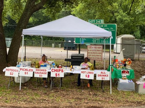 Two vendors under a tent selling hot dogs, pickles, smoked sausage, chips, hot links, and drinks, with prices displayed.