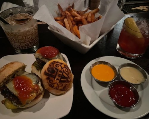 Two mini burgers with pickles and sauces, served with a side of fries and two cocktails on a dark table.