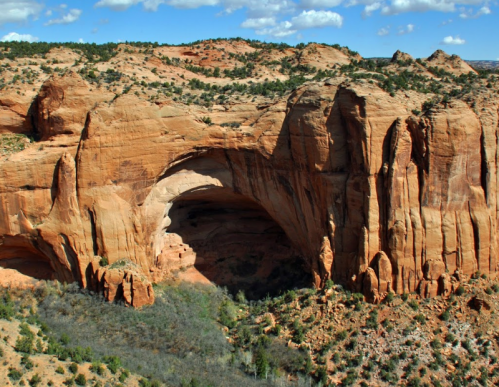 A stunning view of a natural rock arch surrounded by red sandstone cliffs and green vegetation under a blue sky.