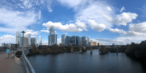 Panoramic view of a city skyline with modern buildings, a river, and a blue sky with fluffy clouds.