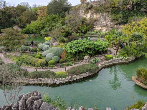 Lush garden with rounded shrubs, a winding path, and a serene green pond surrounded by trees and rocky formations.