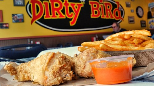 Fried chicken drumsticks with fries and dipping sauce in front of a colorful restaurant sign reading "Dirty Bird."