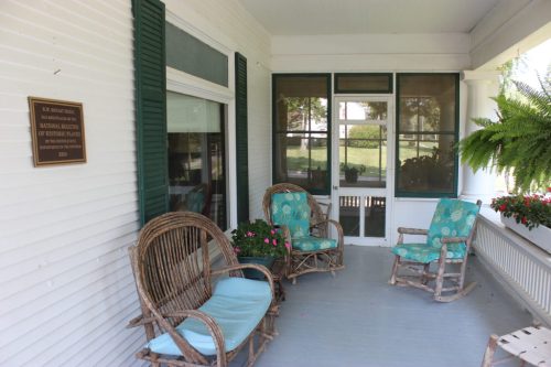 A porch with two rocking chairs, a plaque on the wall, and potted plants, creating a cozy, inviting atmosphere.