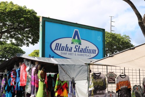 Sign for Aloha Stadium Swap Meet & Marketplace, with colorful clothing stalls in the foreground.