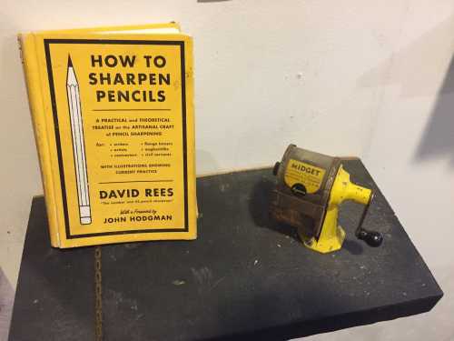 A yellow book titled "How to Sharpen Pencils" next to a vintage pencil sharpener on a display.