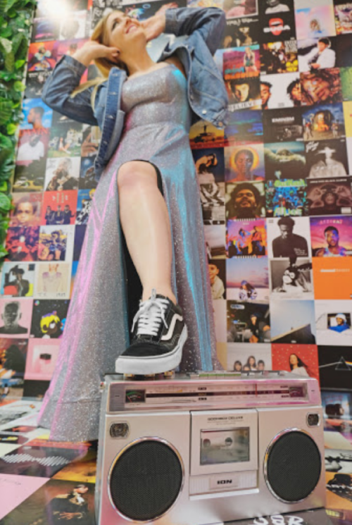 A woman in a sparkly dress poses with a boombox, surrounded by colorful album covers on the wall.