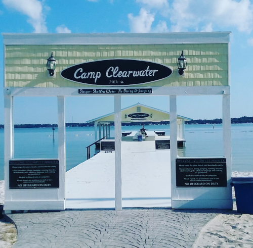 A view of Camp Clearwater pier with a calm water backdrop and clear blue sky. Signs indicate no lifeguard on duty.