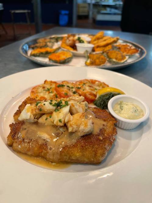 A plate of fried fish topped with sauce and herbs, served with a side of tartar sauce, with appetizers in the background.