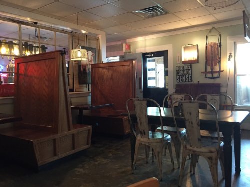 Cozy restaurant interior featuring wooden booths, a communal table, and industrial-style chairs. Warm lighting enhances ambiance.