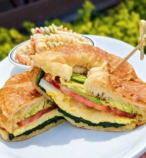 A croissant sandwich filled with spinach, tomato, avocado, and egg, served with a side of pasta salad.
