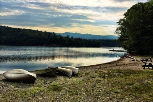 A serene lakeside scene with boats on the shore, surrounded by trees and mountains under a cloudy sky.