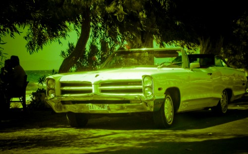 A vintage white convertible car parked under trees, with a beach in the background, tinted in green hues.
