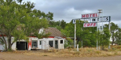 Abandoned motel and café with a weathered sign, surrounded by overgrown grass and trees under a cloudy sky.