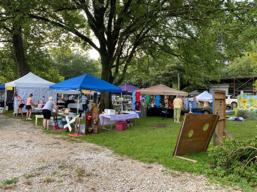 A vibrant outdoor market with colorful tents, vendors, and visitors among trees and greenery.