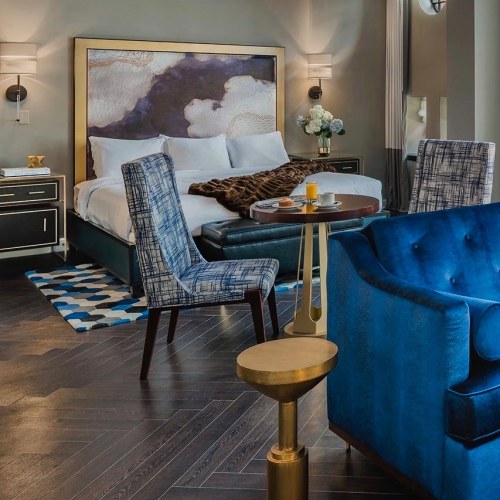 A stylish hotel room featuring a bed, blue chairs, a small table, and modern decor with dark wood flooring.