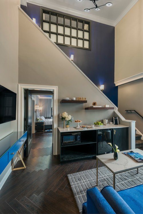 A stylish hotel room with a kitchenette, blue sofa, and staircase leading to an upper level. Modern decor and lighting.