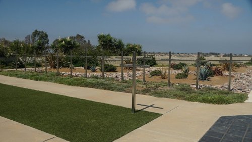 A landscaped area with a cross, featuring plants, rocks, and a pathway under a clear blue sky.