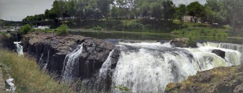 A panoramic view of a waterfall cascading over rocky cliffs, surrounded by greenery and a calm river.