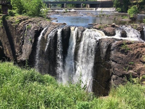 A waterfall cascading over rocky cliffs, surrounded by lush greenery and a river, with a bridge in the background.