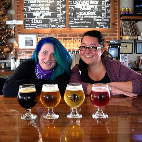 Two women smile at the camera behind a bar, showcasing four glasses of different craft beers in front of them.