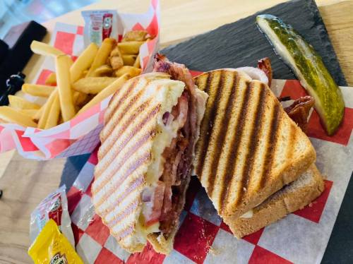 A grilled sandwich with bacon and cheese, served with fries, a pickle, and condiments on a wooden platter.